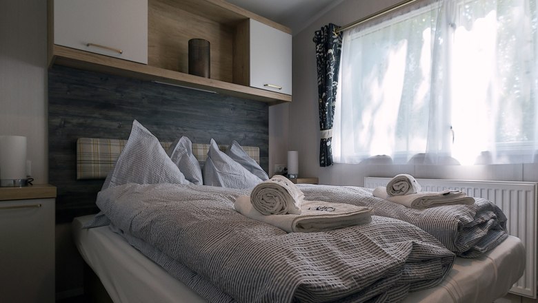 Schlafzimmer, © Leopold Pluschkowitz Ein gemütliches Schlafzimmer mit einem Doppelbett, grauer Bettwäsche und gerollten Handtüchern. Ein Fenster mit Vorhängen lässt Tageslicht herein.