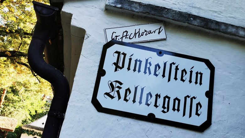 Pinkelstein wine cellar lane in Raschala, © Weinstraße Weinviertel Sign with the inscription 'Pinkelstein Kellergasse' on a white wall.