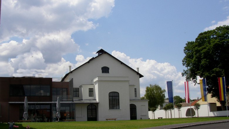 Konzerthaus Weinviertel, © Marktgemeinde Ziersdorf Ein modernes Konzerthaus mit weißen Wänden und großen Fenstern, umgeben von Flaggen und Bäumen, unter einem bewölkten Himmel.