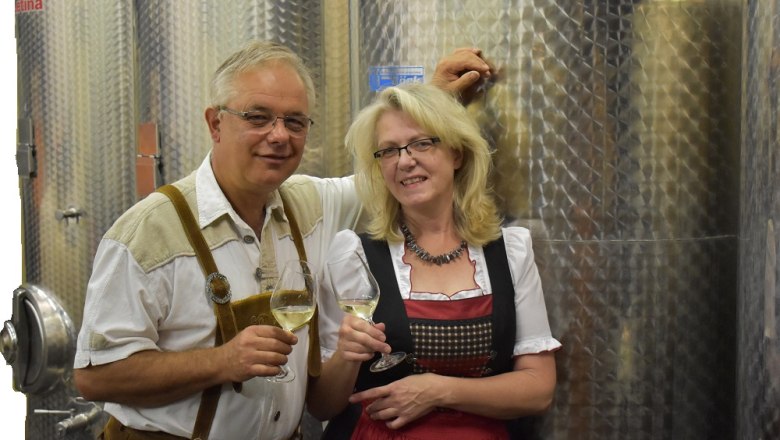 Keller, © Weingut Schober Ein Mann und eine Frau in traditioneller Kleidung stehen vor Weintanks und halten Weingläser.