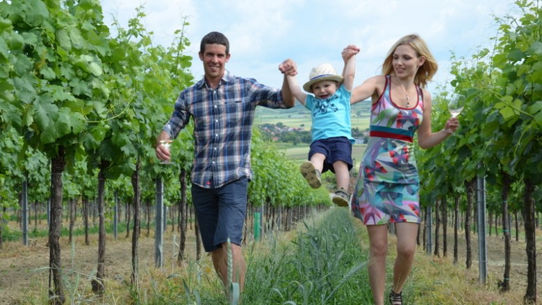 Im Weingarten, © Weingut Markus Lutzer Eine Familie spaziert fröhlich durch einen Weinberg, die Eltern halten ihr Kind an den Händen.