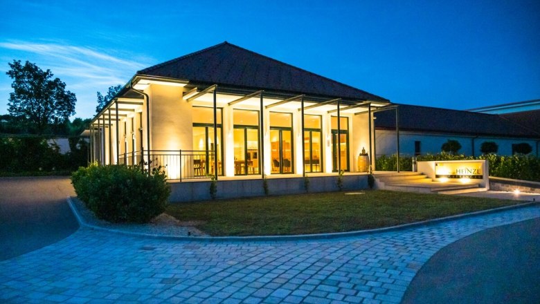 Moderne Architektur und feinste Weine, © Wolfgang Artner Ein modernes Gebäude mit Glasfronten, beleuchtet in der Dämmerung, umgeben von einem gepflasterten Weg und Grünflächen.