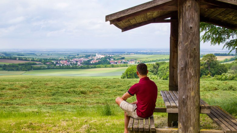 Panoramablick, © Fotostudio Semrad Ein Mann sitzt unter einem Holzunterstand und blickt auf eine weite Landschaft mit Feldern und einem Dorf in der Ferne.
