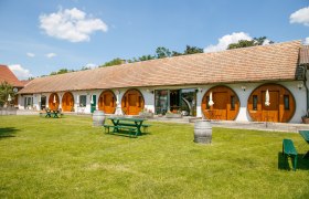 Winzerhof Küssler, © Winzerhof Küssler Ein langes Gebäude mit runden Holztüren und einem gepflegten Rasen davor, auf dem Picknicktische und Weinfässer stehen.