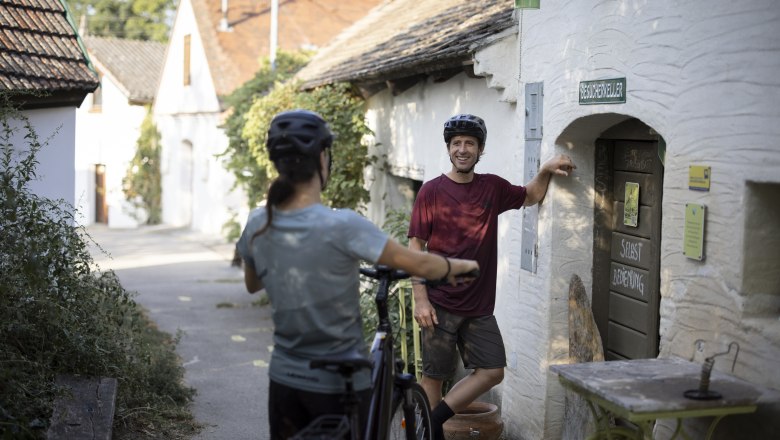 Visitors' cellar on Galgenberg, © Weinviertel Tourismus GmbH / Frühmann Two cyclists in a wine cellar lane, one leaning against a door labeled 'Besucherkeller'.