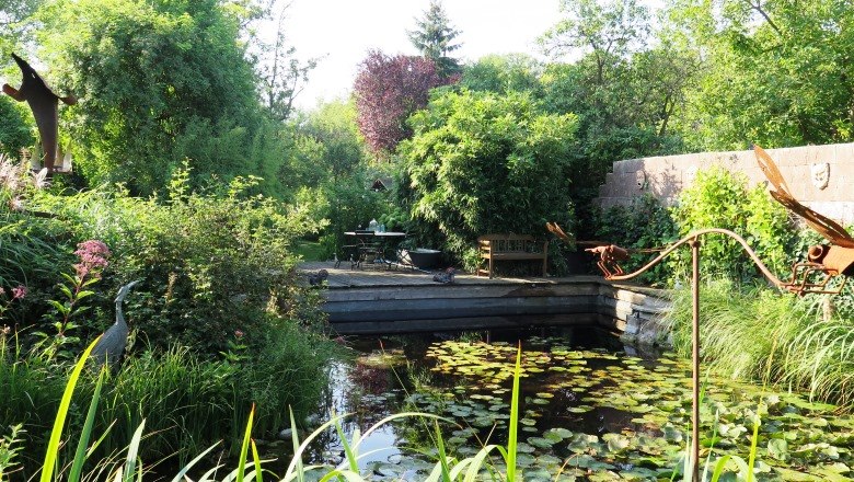 Platz am Teich, © Natur im Garten/Martina Liehl-Rainer Ein idyllischer Garten mit Teich, Seerosen und üppiger Vegetation. Im Hintergrund stehen ein Tisch und Stühle auf einer Terrasse.