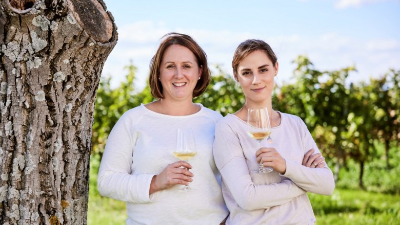 Kerstin und Nadine Schüller, © Stefan Joham Zwei Frauen stehen mit Weingläsern vor einem Baum in einem Weinberg.