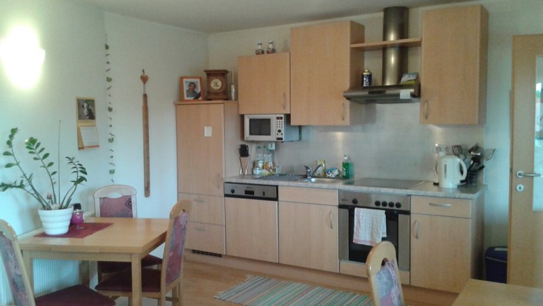 Küche, © © Bianca Waysmayer Eine gemütliche Küche mit Holzmöbeln, Esstisch, Stühlen, Spüle, Herd und Mikrowelle. An der Wand hängen Bilder und ein Kalender.