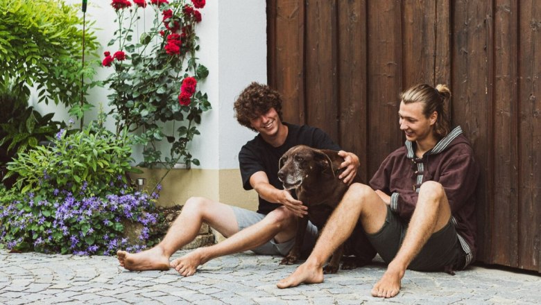 Florian und Lorenz Widerna, © Elke Wolfbeisser Zwei Männer sitzen barfuß auf einem gepflasterten Boden neben einem braunen Hund. Im Hintergrund sind rote Rosen und eine Holzwand zu sehen.