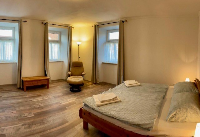 Schlafzimmer, © Julia Grünschachner Ein schlichtes Schlafzimmer mit Doppelbett, Sessel, Nachttisch und drei Fenstern mit Vorhängen.