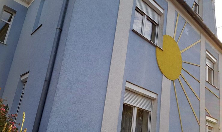 Hausansicht, © Harald Mader Blaues Haus mit gelber Sonnendekoration an der Fassade.