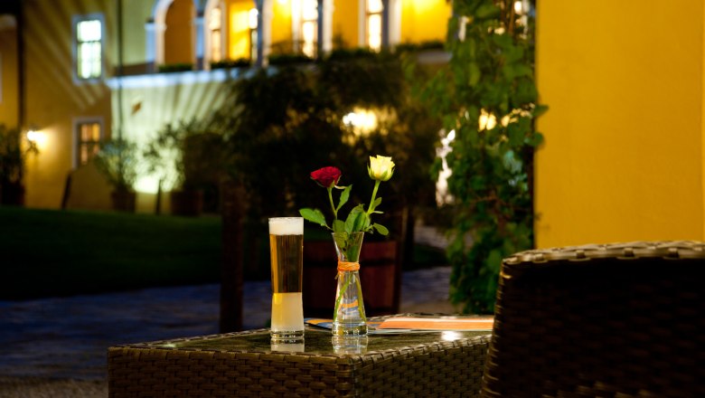 Evening atmosphere in the Althof, © Althof Retz A table with a glass of beer and two roses in a vase, in the background an illuminated building with arches.