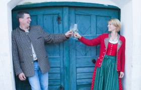 Familie Schönhofer, © Sabine Schneider Ein Mann und eine Frau stoßen mit Weingläsern an, vor einer blauen Holztür.