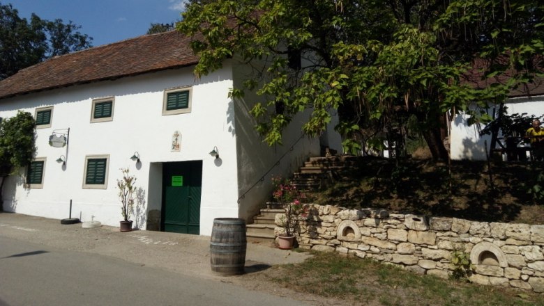 Weingut Himmelbauer, © Weinstraße Weinviertel Weißes Gebäude mit grünen Fensterläden und offener Tür, umgeben von Bäumen und einem Weinfass auf der Straße.