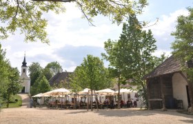 Dorfwirtshaus im Museumsdorf, © Museumsdorf / Bodensteiner Ein Dorfwirtshaus im Museumsdorf mit Sonnenschirmen und einer Kirche im Hintergrund.