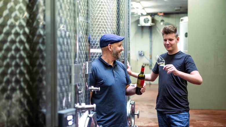 Father and son at work, © Astrid Bartl Two men in a wine cellar, one holding a wine bottle, the other a wine glass.