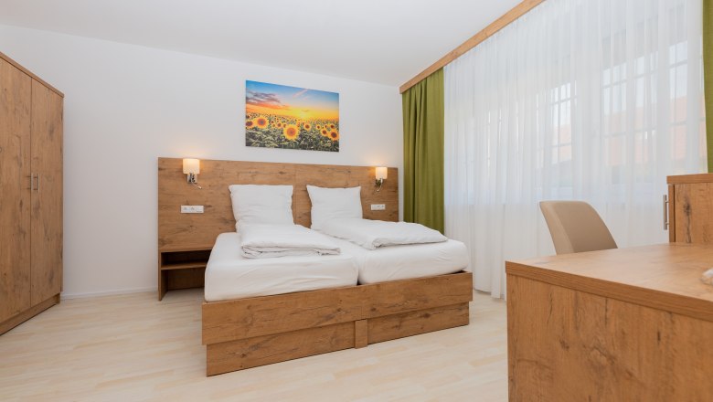 Schlafzimmer, © Schönfelderhof Modernes Schlafzimmer mit Holzmöbeln, Doppelbett, Schrank und Schreibtisch. Ein Bild mit Sonnenblumen hängt über dem Bett.