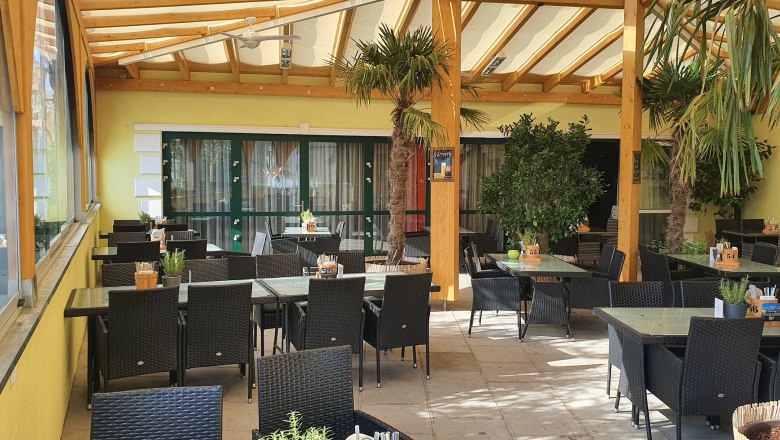Restaurant Lebenszeit, © Familie Eissler Überdachte Terrasse eines Restaurants mit schwarzen Rattanmöbeln und Pflanzen.