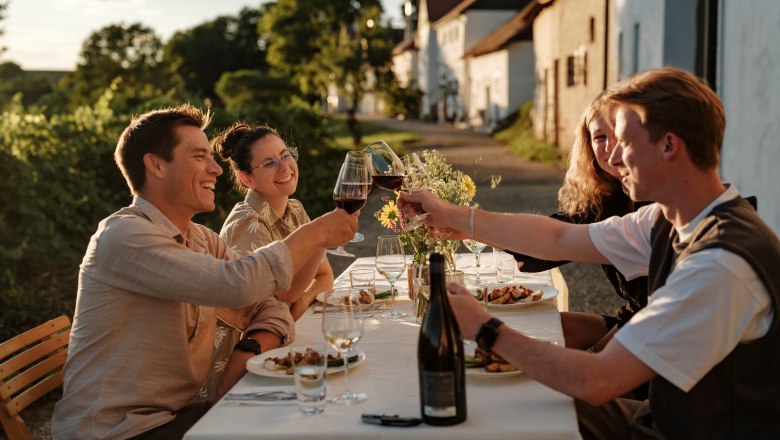 Weinviertler Kellergassenkulinarium, © Weinstraße Weinviertel / Reidinger Eine Gruppe von Menschen stößt bei einem Abendessen im Freien mit Weingläsern an. Im Hintergrund sind Weinberge und Häuser zu sehen.