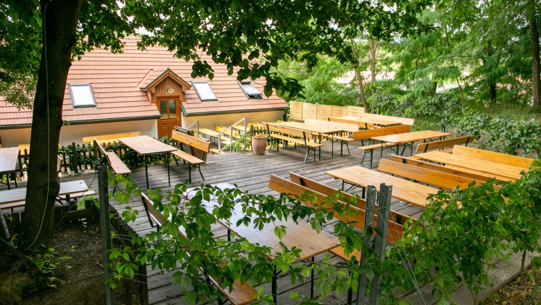 Winery Küssler, © Winzerhof Küssler An empty beer garden with wooden benches and tables under trees, next to a building with a red tiled roof.