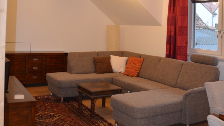 Wohnzimmer, © Familie Herrmann Modernes Wohnzimmer mit grauem Ecksofa, Holzbalken, Teppich und Lampe.