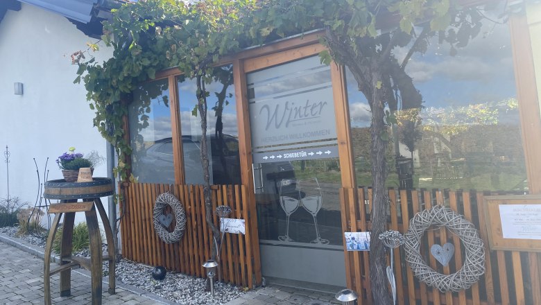 Winter Winery, © Cathy Winter Entrance of a building with wooden paneling and vines, decorated with wreaths and a table with flowers.