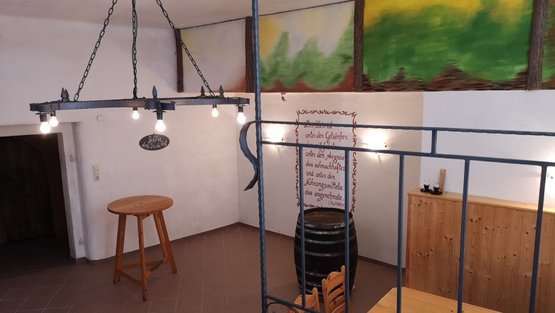 Bauers Kellerrast, © Weinbau Bauer Innenansicht eines rustikalen Kellers mit Holztisch, Weinfass und Wanddekoration.