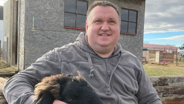 An experience for the whole family, © Sabine Valis A man is sitting outside next to a black sheep, which he is stroking. A small building can be seen in the background.
