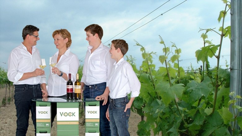Familie Vock, © Franz Vock Vier Personen stehen in einem Weinberg und stoßen mit Weingläsern an. Vor ihnen ein Tisch aus Vock-Weinkartons.