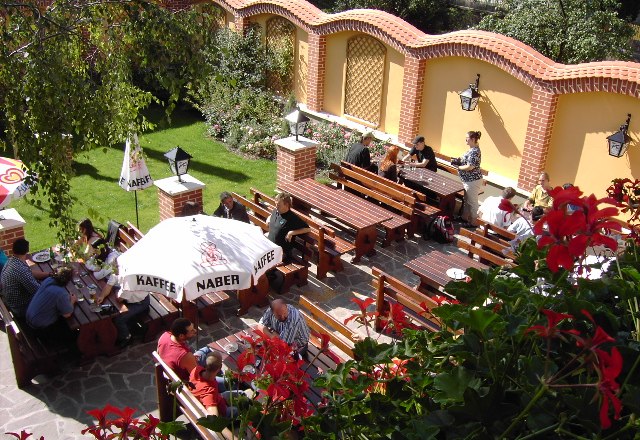 Gastgarten, © Stefanshof Ein sonniger Gastgarten mit Holzbänken und Tischen, umgeben von einer gelben Mauer und Pflanzen.