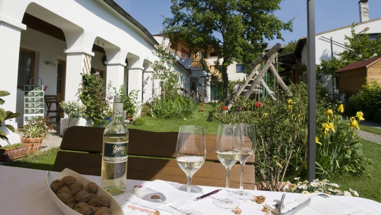 Inner courtyard, © Michael Himml An idyllic courtyard with a table, wine bottle, glasses and nuts in the foreground. In the background, a garden with flowers and a tree.