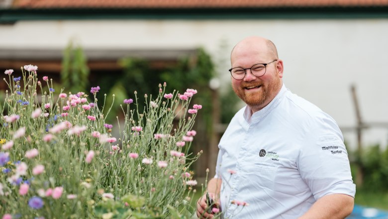 Herbst Inn, © Weinviertel Tourismus / Michael Reidinger A smiling man in chef's clothes stands next to blooming flowers outside.
