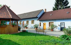 Weinbauernhof Kahrer, © Fam. Kahrer Ein idyllischer Hof mit einem Pavillon und einem blauen Gebäude mit roten Dächern, umgeben von Pflanzen.