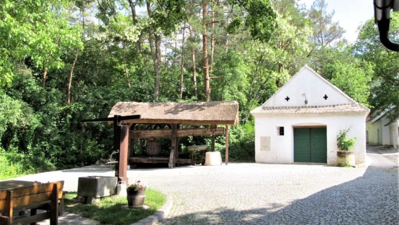 Weinvierlter Kellergassenkulinarium, © D`Raschalaer Köllamauna Eine gepflasterte Kellergasse mit einem kleinen weißen Gebäude und einer überdachten Holzpresse, umgeben von Bäumen.