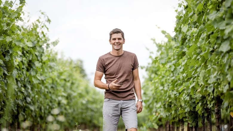 Winzer Roman Degn, © Astrid Bartl Ein Mann in einem Weinberg, lächelnd und in Freizeitkleidung, umgeben von grünen Weinreben.