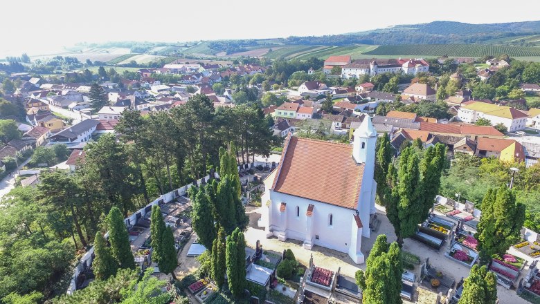Kunigundenkirche with surrounding cemetery, © Marktgemeinde Mailberg Aerial view of a church with a red roof, surrounded by a cemetery and trees, in a rural setting with houses and hills in the background.