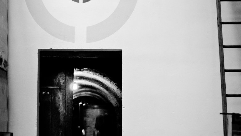 Obenaus-Keller, © Bernhard Kastner Ein schwarz-weiß Foto eines Kellers mit einem Stuhl vor einer Tür und einem Zielscheibenmuster an der Wand.