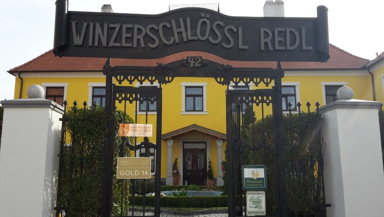 Winzerschlössl, © Weingut Redl Eingangstor zum Winzerschlössl Redl mit gelbem Gebäude im Hintergrund.