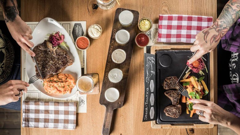 Ignition unit, © Lukas Kirchgasser Photography Two people eat steaks with side dishes at a wooden table.