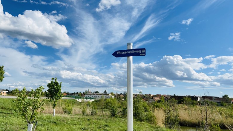 Spaziergang entlang des Wasserweltweges, © Weinstraße Weinviertel Ein Wegweiser mit der Aufschrift 'Wasserweltweg' steht auf einer grünen Wiese unter einem blauen Himmel mit weißen Wolken.