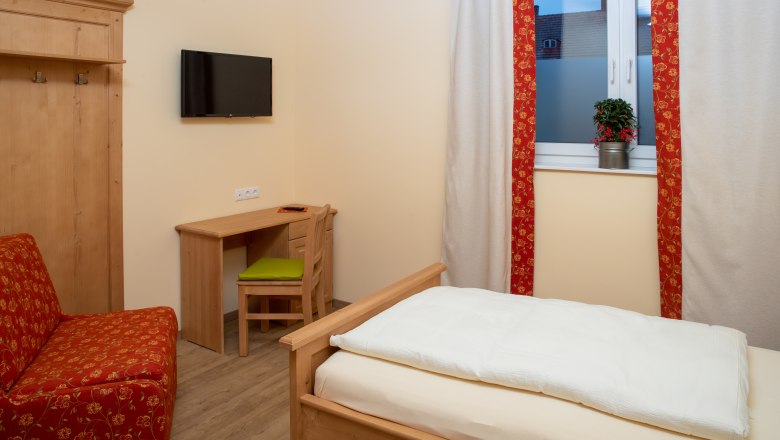 Einzelzimmer, © Nadine Beles Ein gemütliches Einzelzimmer mit Holzmöbeln, einem Bett, einem Schreibtisch und einem Fernseher an der Wand. Ein Fenster mit Vorhängen und einer Pflanze.