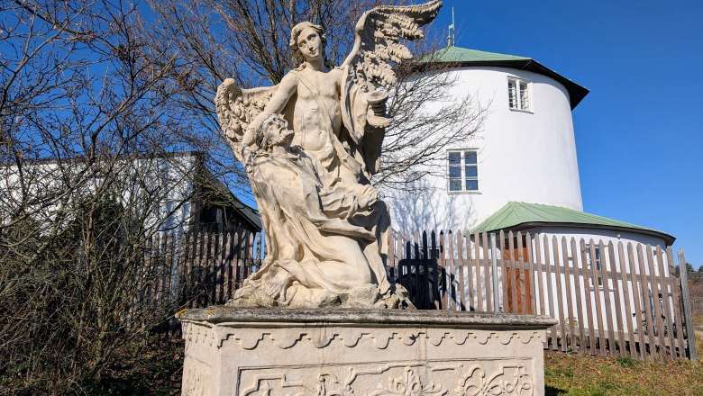 Kalvarienberg, © Retzer Land / Daniel Wöhrer Steinstatue eines Engels mit Flügeln und einer knienden Figur vor einem runden, weißen Gebäude mit grünem Dach.