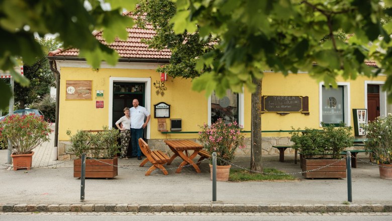 Herbst Inn, © Weinviertel Tourismus / Michael Reidinger A yellow inn with a red roof, surrounded by trees. Two people are standing in front of the entrance.