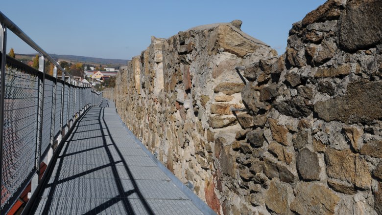Rundgang auf Stadtmauer, © Veigl Harald Ein Metallsteg entlang einer alten Steinmauer mit Blick auf eine Stadt im Hintergrund.