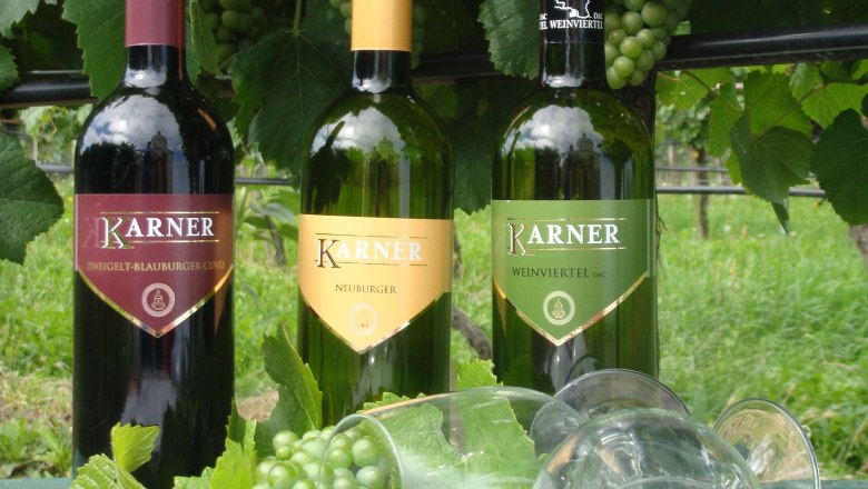 Weingut Karner, © Weingut Karner Drei Weinflaschen von Karner mit Trauben und Weingläsern auf einem Tisch vor Weinreben.