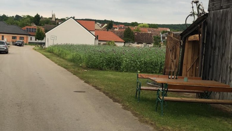 Radlerrast Pottenhofen, © Lukas Cermak Dorfstraße mit Holzhütte und Fahrrad auf dem Dach, Bänke und Tisch daneben, Felder und Häuser im Hintergrund.