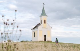 Michelberg am Jakobsweg Weinviertel, © Weinviertel Tourismus / Claudia Schlager Eine kleine Kapelle auf einem Hügel mit einer Person, die darauf zugeht, umgeben von einer Wiese und bewölktem Himmel.