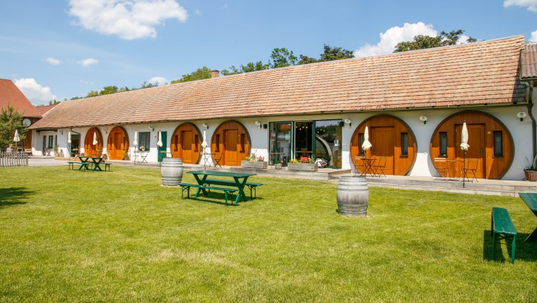 Winzerhof Küssler, © Winzerhof Küssler Ein langes Gebäude mit runden Holztüren und einem gepflegten Rasen davor, auf dem Picknicktische und Weinfässer stehen.