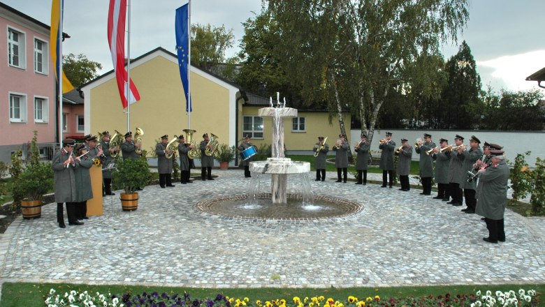 Enzersfeld, © Gemeinde Enzersfeld Eine Blaskapelle spielt im Freien vor einem Brunnen, umgeben von Blumen und Flaggen.