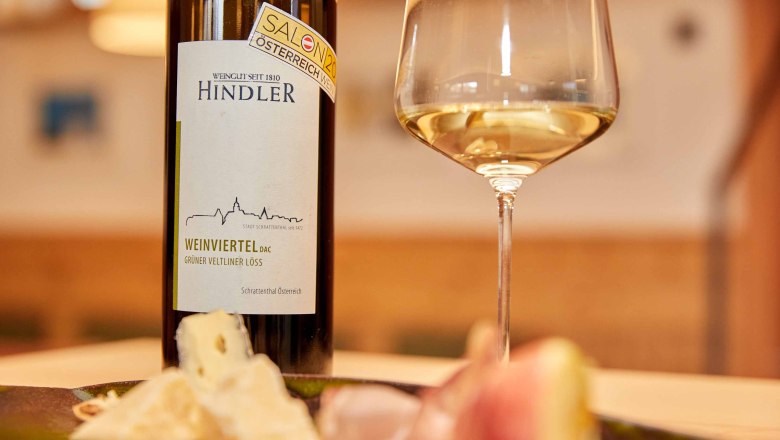 Weingut Hindler, © Weingut Hindler Weinflasche und Glas mit Weißwein, Käse und Schinken im Vordergrund.